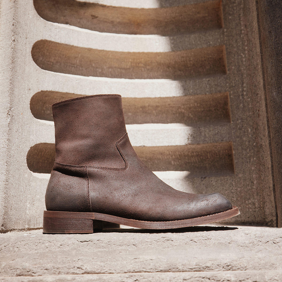 A brown ankle boot with a low block heel, shown in profile against a textured concrete wall with horizontal wooden slats casting shadows.