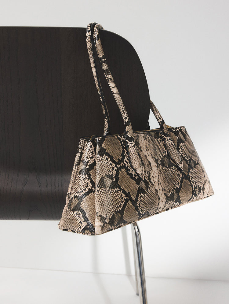 A snakeskin-patterned shoulder bag with beige, brown, and black tones, hanging from the back of a black chair against a white studio background.