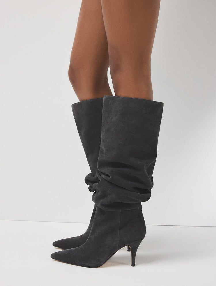 A pair of dark gray suede knee-high boots with pointed toes and kitten heels, shown on a model's feet against a light gray studio background.