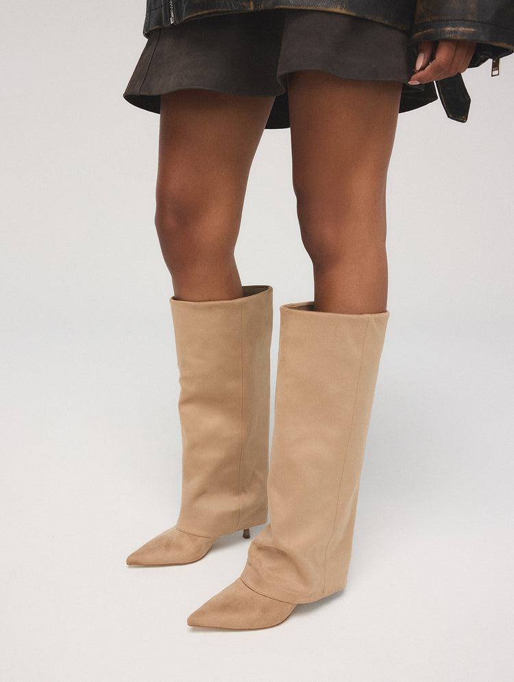 Une personne portant une paire de hautes bottes beige à hauteur du genou, à bout pointu et à talon bloc bas, photographiée à partir des genoux vers le bas sur un fond de studio gris pâle.