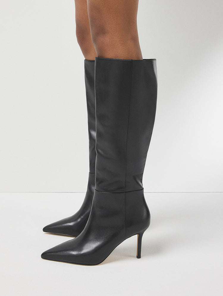 Une paire de bottes hautes en cuir noir à bout pointu et à talons aiguilles fins, portées par un mannequin et photographiées sur un fond de studio gris pâle.