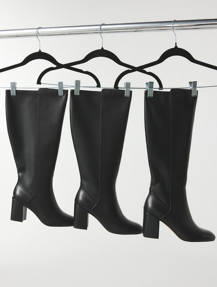 Trois bottes hautes en cuir noir à talons blocs suspendues à des cintres noirs sur un portant à vêtements, devant un fond gris pâle.