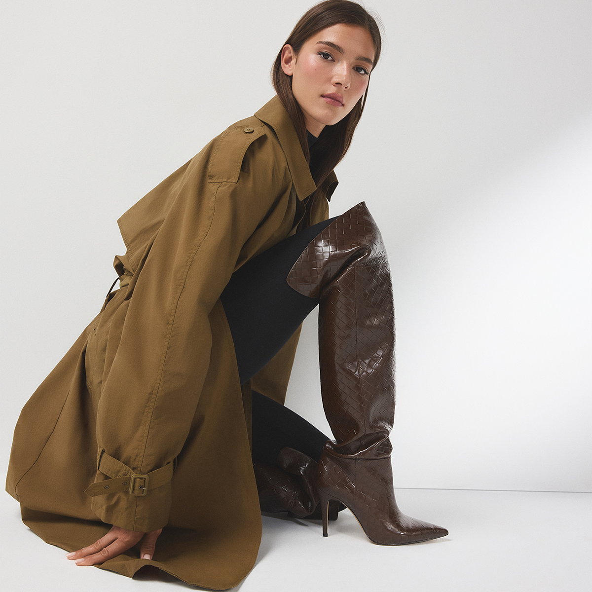 Un mannequin agenouillé sur un plancher de studio gris pâle, portant un trench-coat brun surdimensionné, des collants noirs et une botte haute brun foncé lustrée à bout pointu et à talon, sur un fond de studio gris pâle.