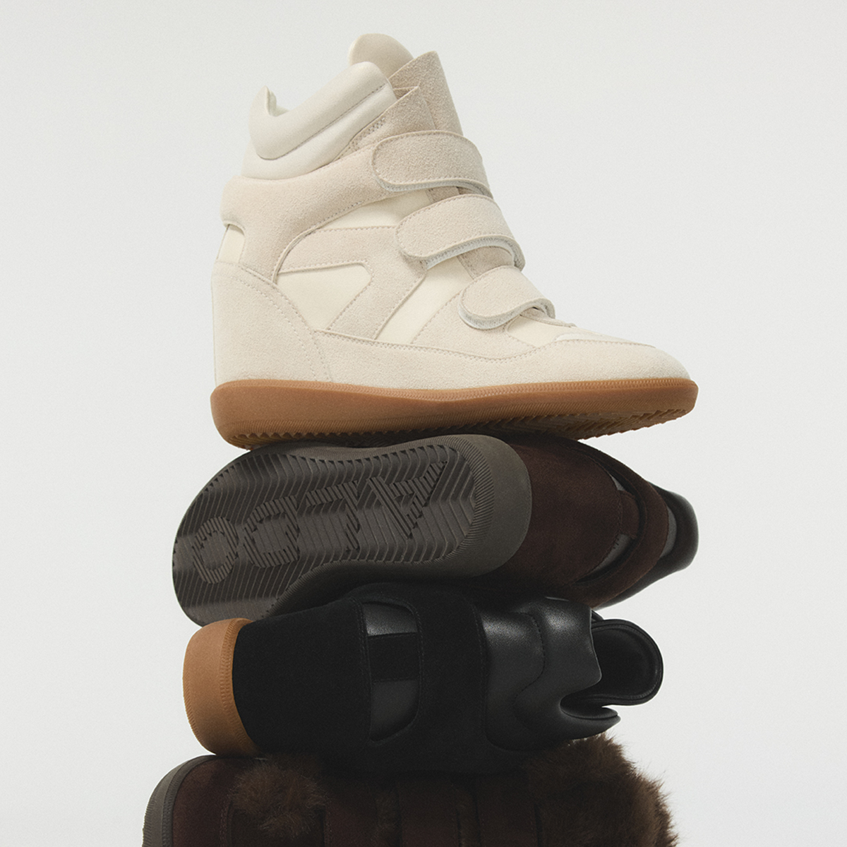 Une espadrille montante de couleur crème munie de trois brides à fermeture autoagrippante et d’une semelle couleur gomme, empilée sur plusieurs autres chaussures dans des tons de noir, de brun et de suède, devant un fond gris pâle uni.