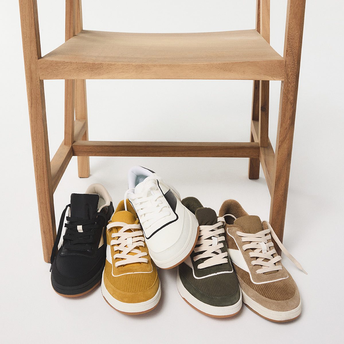Five pairs of men's Ashwin sneakers in different colors arranged on a white floor with a wooden chair in the background.