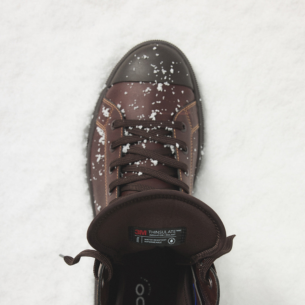 A single brown leather winter boot with a dark brown rubber toe cap, shown from a top-down view on a white snowy surface with light snowflakes dusting the upper.