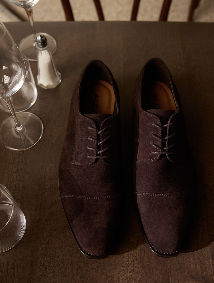 Une paire de souliers oxford en suède brun foncé avec des lacets assortis, présentée sur une table en bois foncé, entourée de verres à vin et d’une salière en arrière-plan.