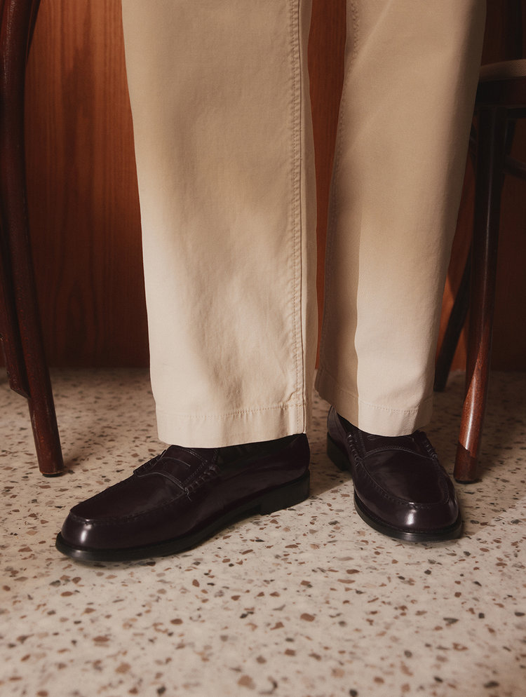 Une vue rapprochée des jambes d’une personne portant un pantalon beige et des mocassins penny en cuir brun foncé, debout sur un plancher en terrazzo moucheté entre des chaises en bois.