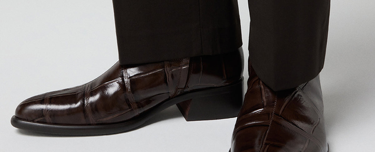 A close-up of a person's feet wearing dark brown patent leather boots with a low block heel and black trousers, shown against a light gray studio background.
