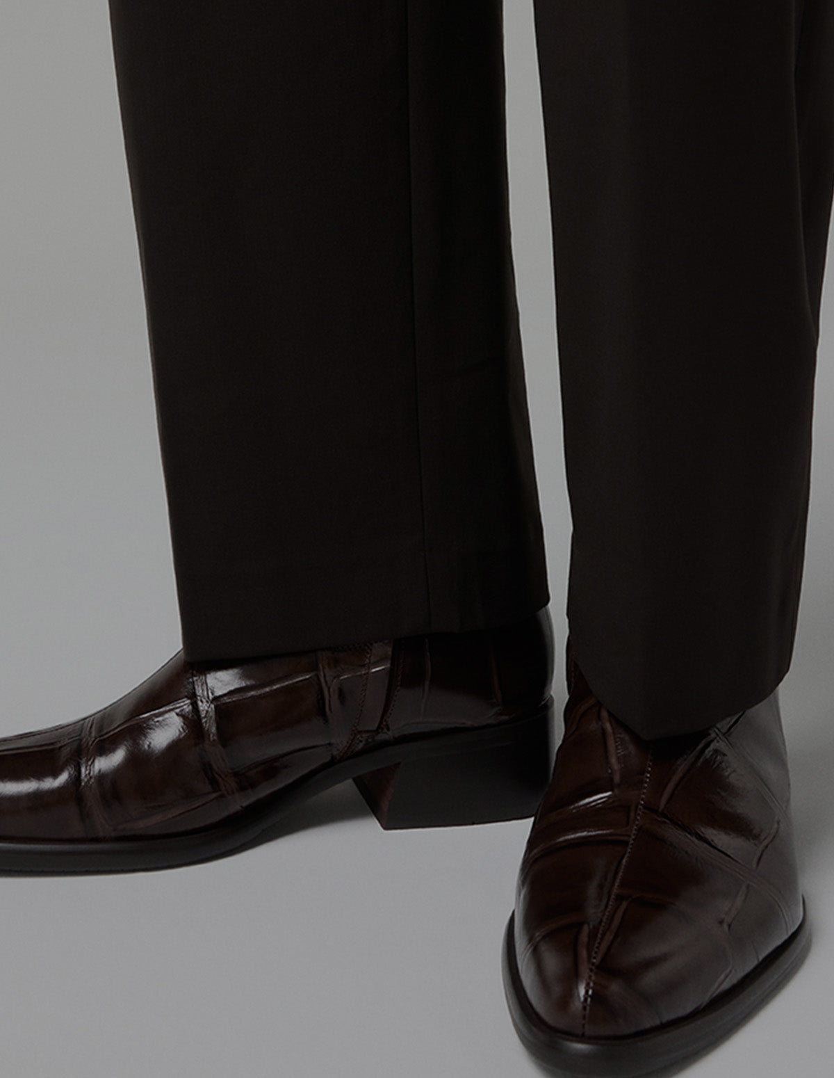 A close-up of a person's feet wearing dark brown patent leather boots with a low block heel and black trousers, shown against a light gray studio background.