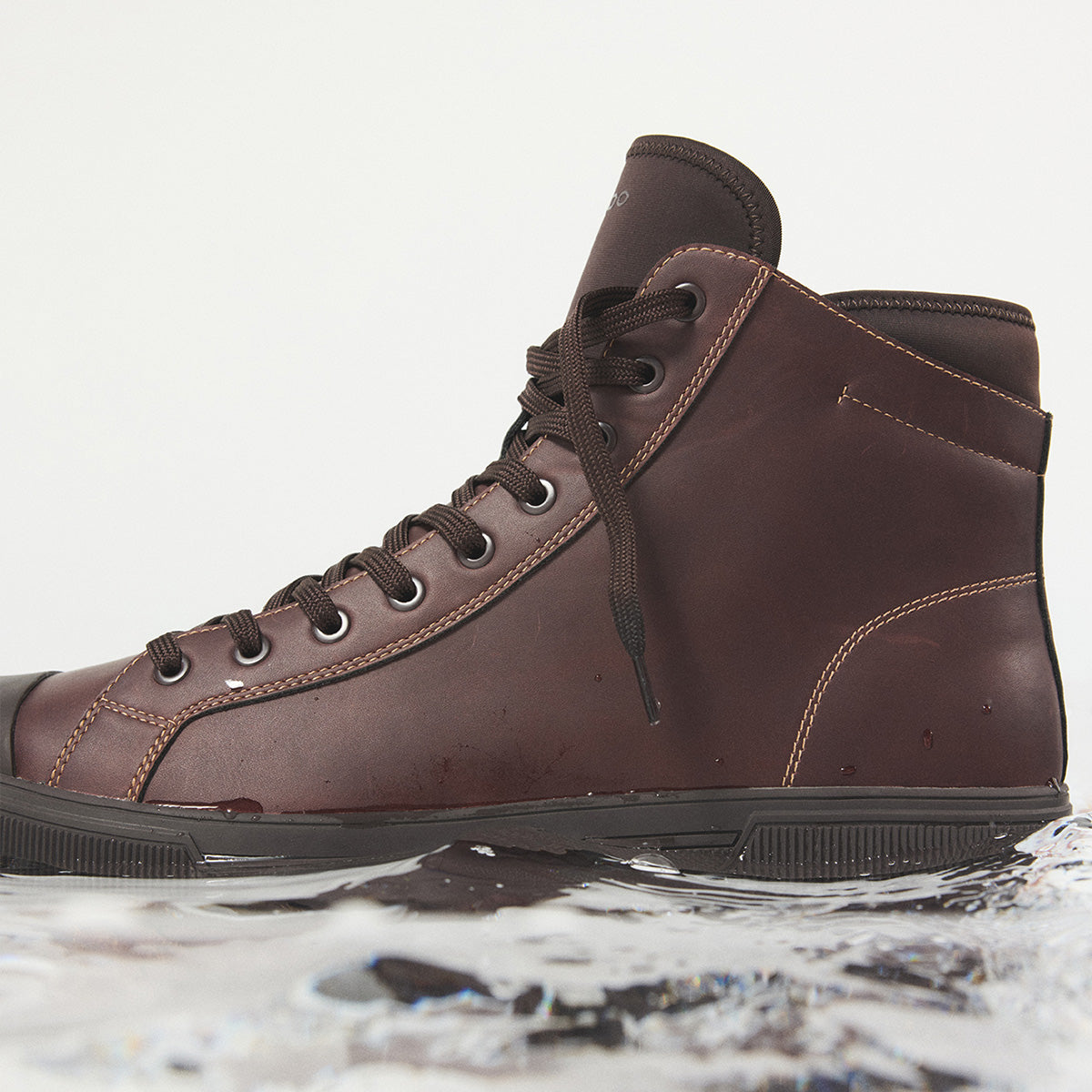 A dark brown leather high-top sneaker boot with dark laces and a black sole, shown resting on a reflective puddle of water against a light gray background.
