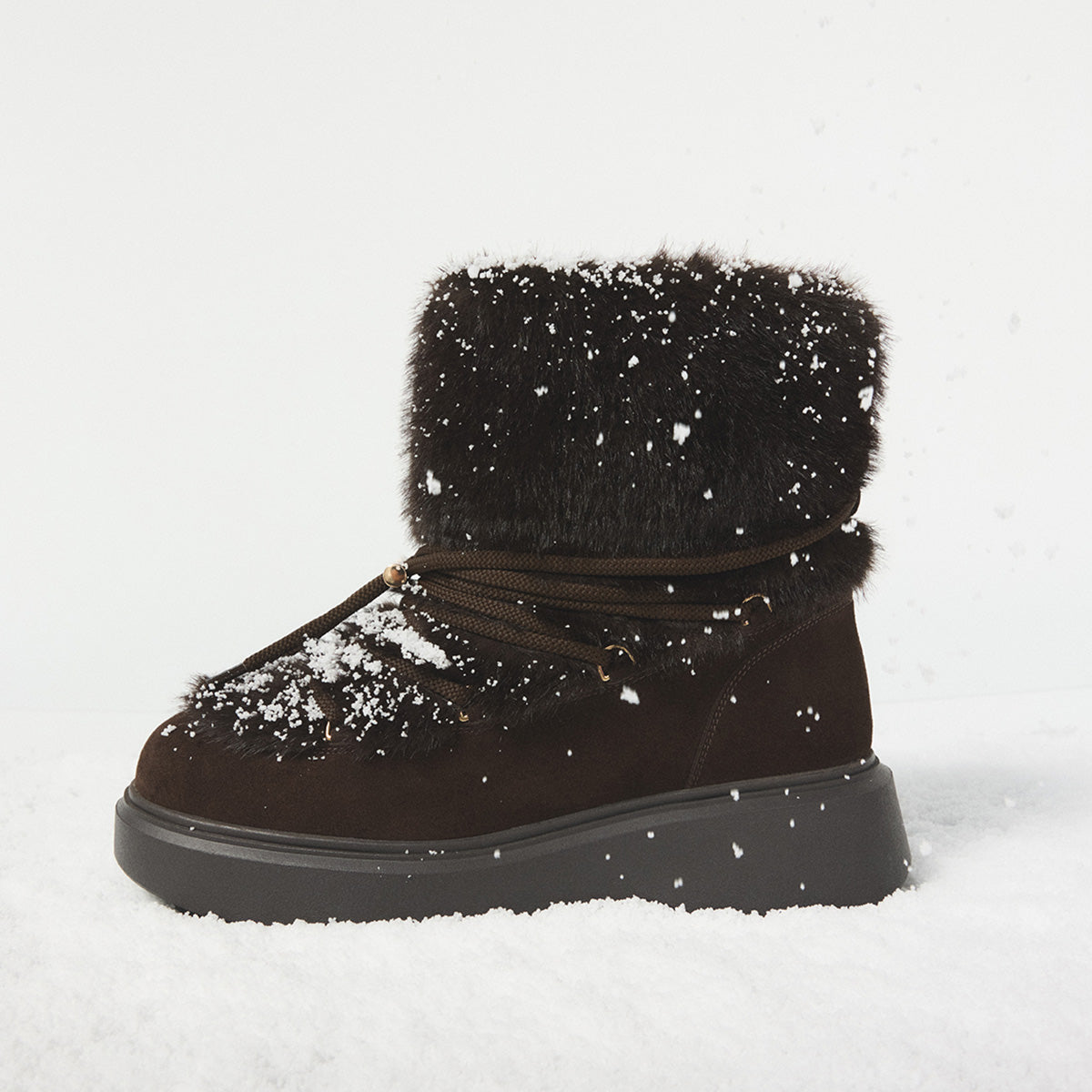 A dark brown suede winter boot with a fluffy black faux-fur collar, resting on white snow with light snowflakes falling. The boot has a thick black sole and brown laces.