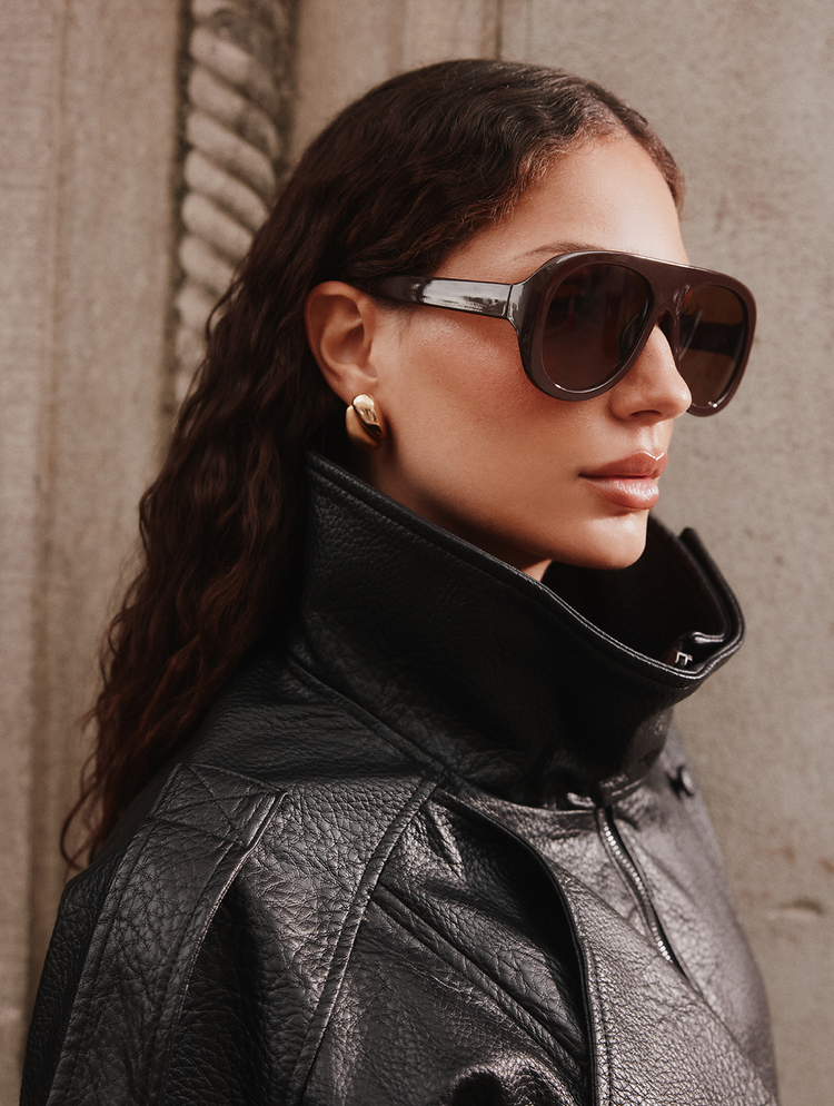 Une femme brune photographiée de profil, portant de grandes lunettes de soleil brunes, des boucles d’oreilles dorées et une veste en cuir noir.