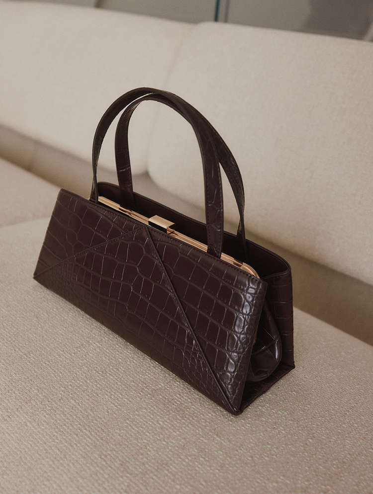 Une sac à main en cuir brun foncé à effet crocodile embossé, doté de deux poignées supérieures et d’un fermoir de couleur or, reposant sur un canapé beige texturé.