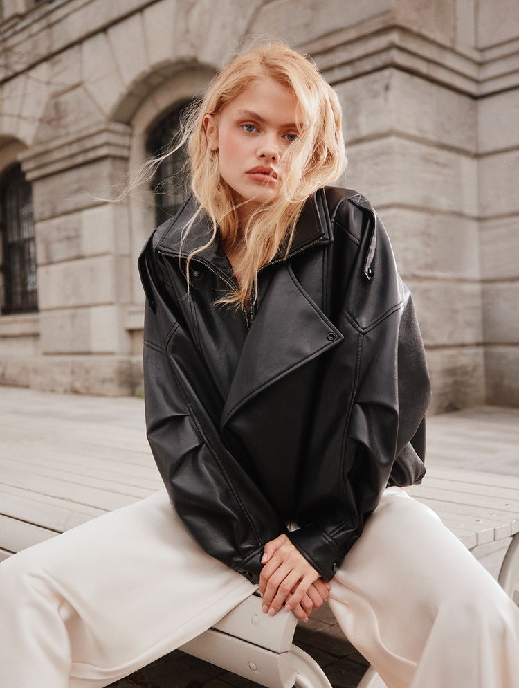 Une femme blonde assise sur un banc, portant une veste en cuir noir et un pantalon blanc.