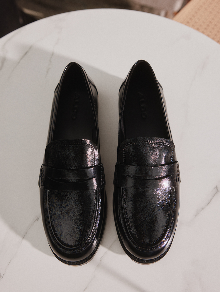 Une paire de mocassins penny en cuir noir au fini lustré, présentée sur une surface en marbre blanc.