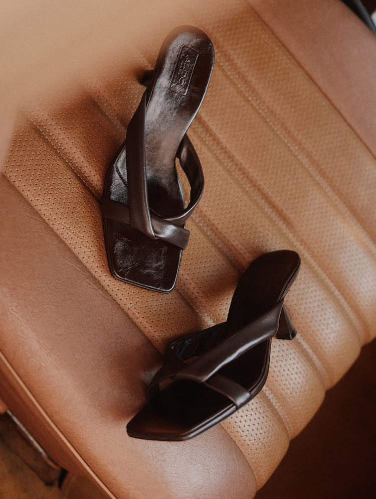 A pair of dark brown leather slide sandals with square toes and low block heels, resting on a tan perforated leather seat.