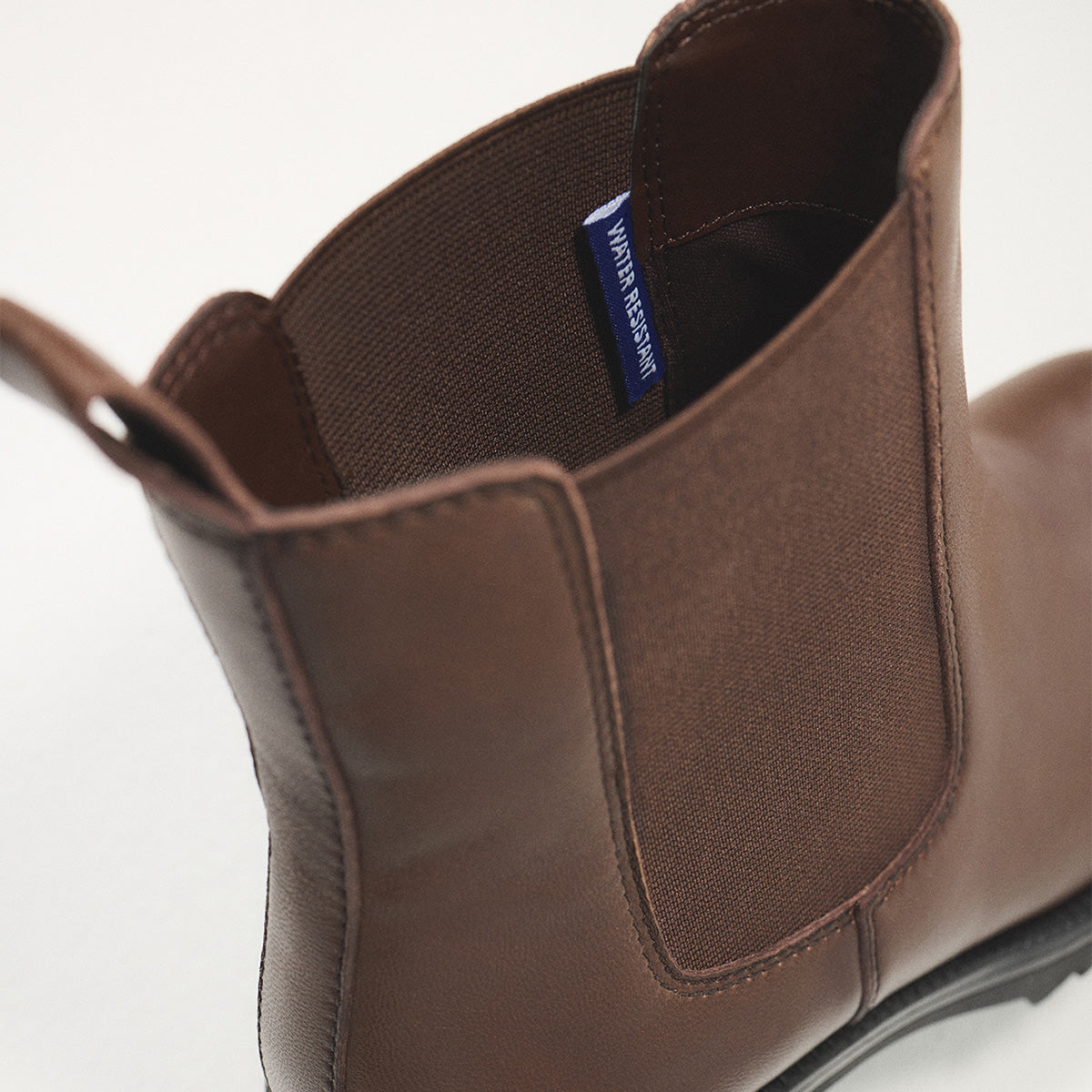 A close-up shot of a brown leather Chelsea boot, showing the side panel, elastic gore, pull tab, and interior lining with a blue 'WATER RESISTANT' brand tag.
