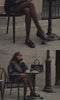 A women sitting outside of a cafe talking on the phone wearing a black jacket, black loafers, and black handbag.