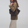 A video of a person wearing a light brown suede jacket, a white silk maxi skirt, and brown mule heels while carrying a khaki suede textured tote bag. It features close-up shots highlighting the bag’s texture and gold-tone hardware, and concludes with the person walking out of the frame carrying the bag.