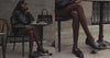 A women sitting outside of a cafe talking on the phone wearing a black jacket, black loafers, and black handbag.