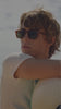 A video of a man with sunglasses throwing a beach ball, a close of sandals on the beach and two people sitting on the sand.