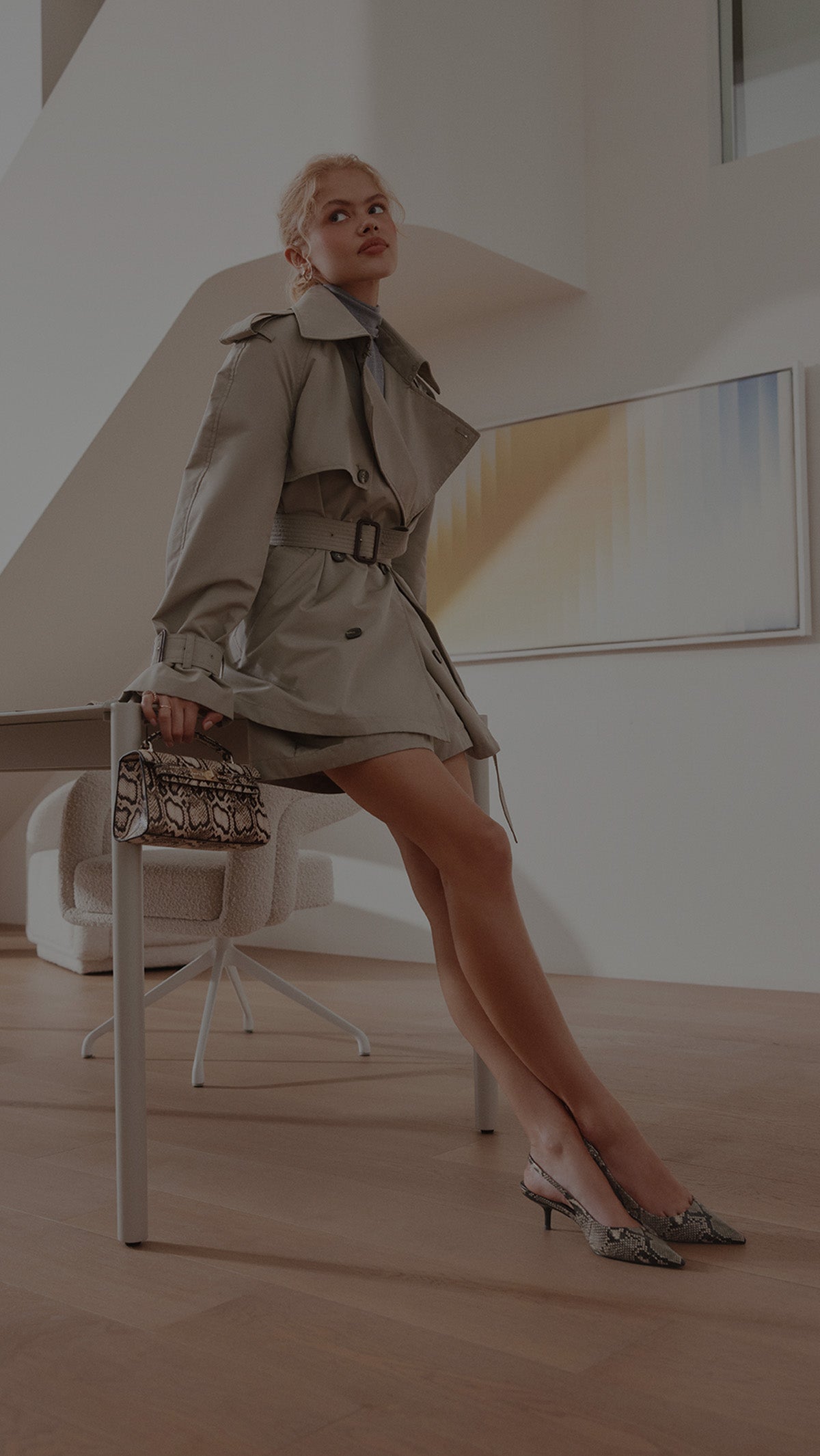 A woman posing in a beige trench coat and matching mini skirt, holding a snakeskin-patterned handbag, in a modern minimalist room with light wood floors.