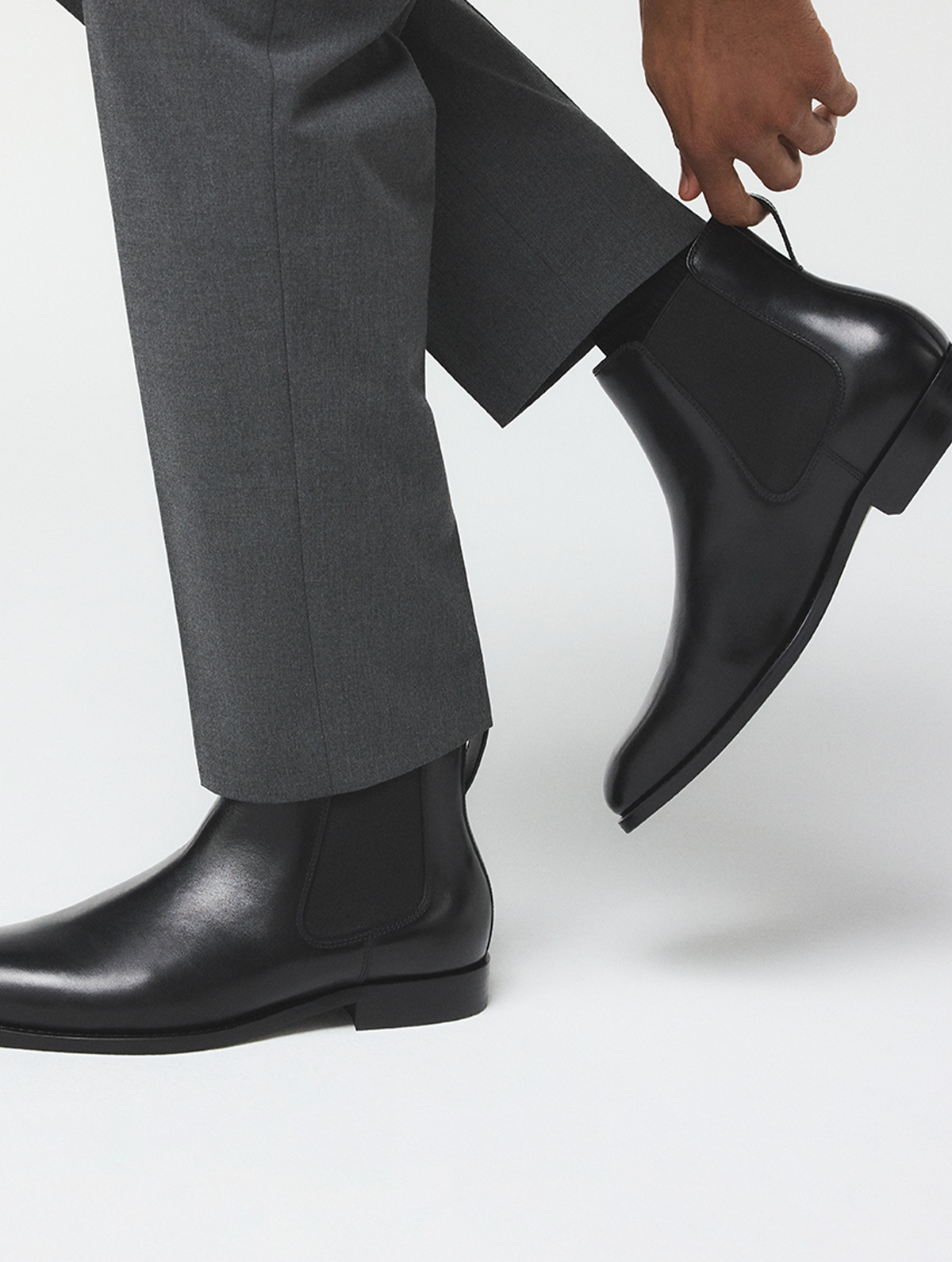 Une vue rapprochée des pieds d’une personne portant des bottines Chelsea noires avec un pantalon gris foncé, soulevant l’un des bottillons pour en mettre en valeur le profil latéral, sur un fond de studio blanc épuré.