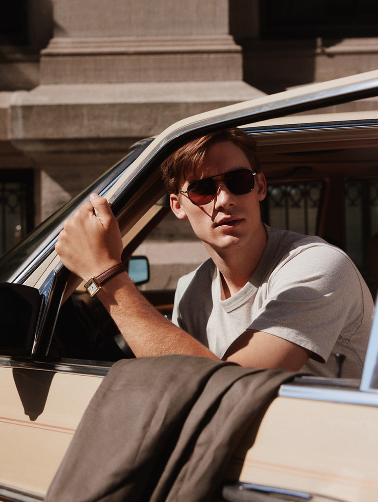 Un homme portant des lunettes de soleil de style aviateur foncées et un t-shirt gris pâle se penche à la fenêtre d’une voiture vintage de couleur crème dans une rue ensoleillée.