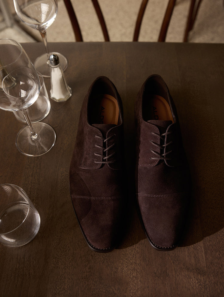 Une paire de souliers derby en suède brun foncé présentée sur une table en bois foncé, entourée de verres à vin et d’une salière.