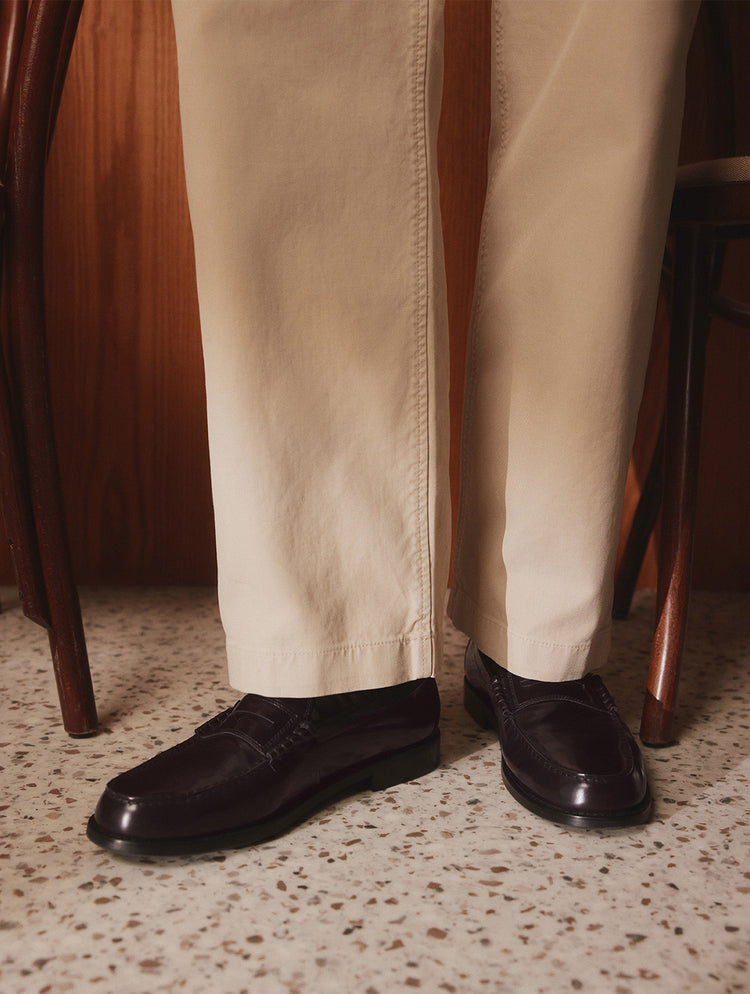 Une vue rapprochée d’une personne portant un pantalon beige et des mocassins penny en cuir brun foncé, debout sur un plancher en terrazzo moucheté entre des chaises en bois.