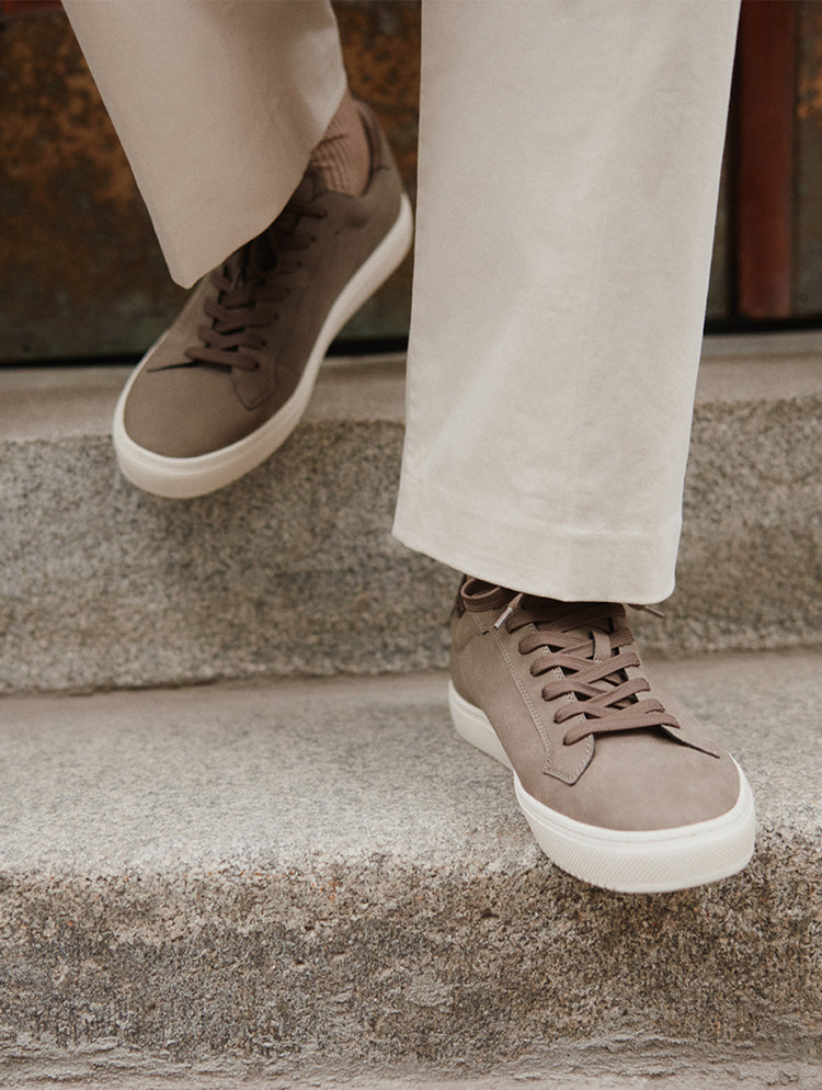 Une personne portant des espadrilles en suède brun taupe à semelles blanches, agencées à un pantalon beige pâle, descendant des marches en béton.
