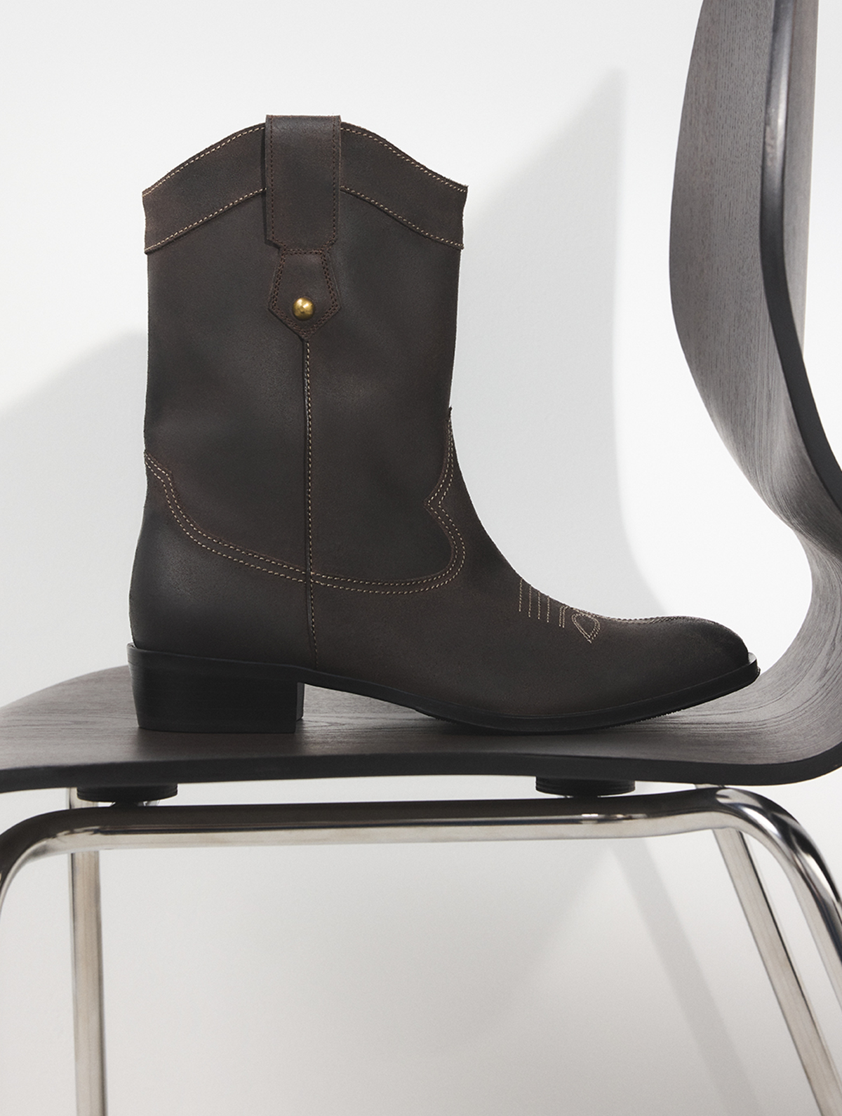 Une botte de cowboy en cuir brun foncé à talon bloc bas et à surpiqûres décoratives, présentée sur une chaise noire moderne devant un fond blanc.