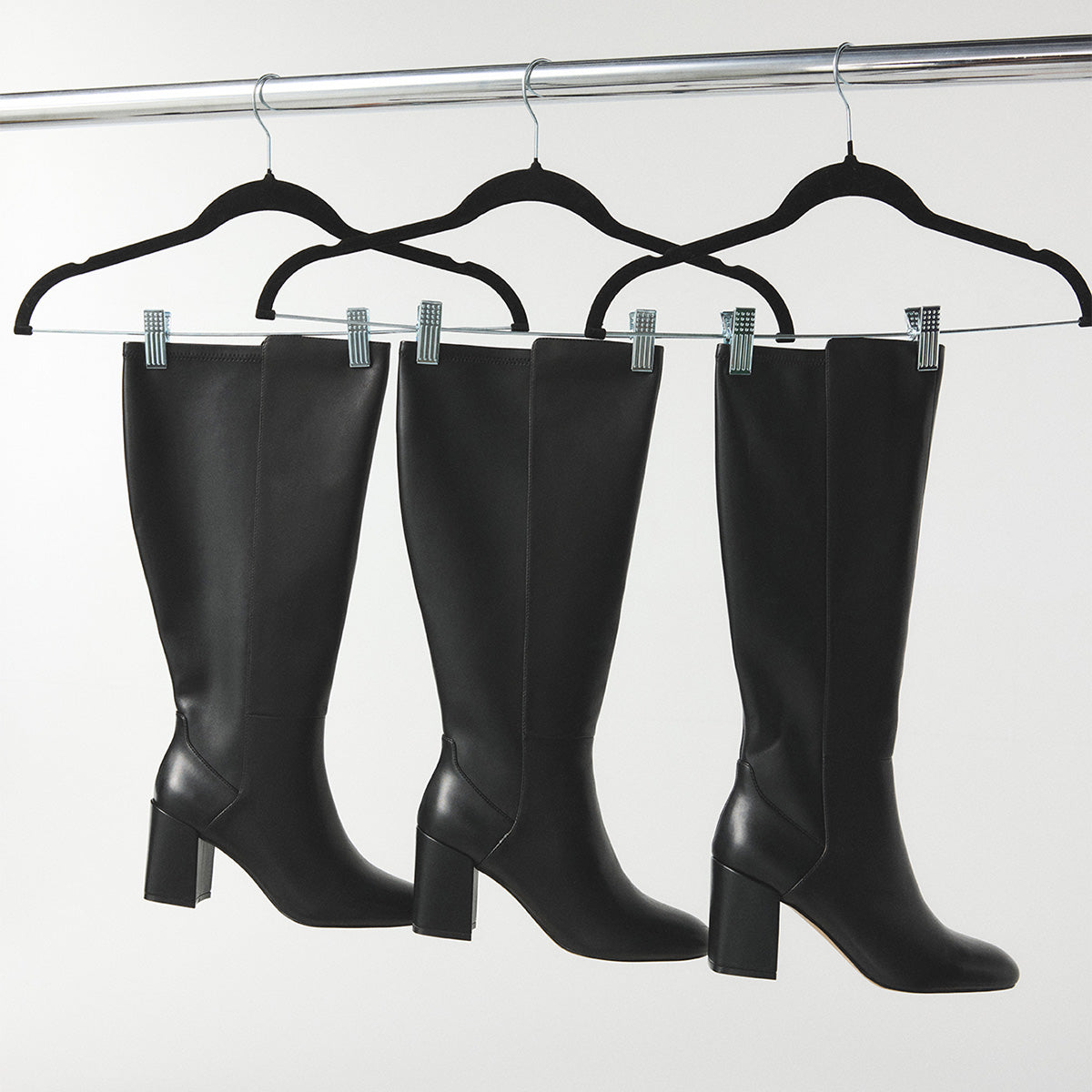 Four black knee-high leather boots with block heels hanging from black hangers on a silver clothing rack against a light gray background.