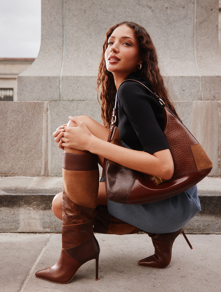Une femme accroupie sur des marches en pierre à l’extérieur, portant des bottes hautes en cuir brun à talons, un haut noir à manches courtes et une jupe bleue, tout en transportant un grand sac porté à l’épaule en cuir brun.