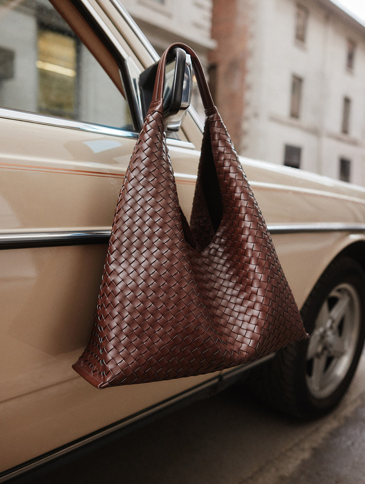 A brown woven leather shoulder bag hanging from a car window, with a beige car and blurred city buildings in the background.