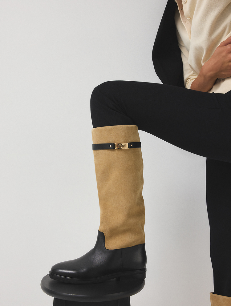 La jambe d’un mannequin est allongée, portant une haute botte en suède tan avec une partie inférieure en cuir noir et une bride en cuir noir ornée d’une boucle de couleur or, agencée à un pantalon noir et une chemise crème.