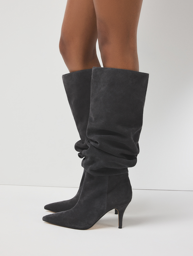 Une paire de bottes hautes en suède gris foncé à effet plissé, à bout pointu et à talon de type kitten heel, portées par un mannequin et photographiées sur un fond de studio blanc épuré.