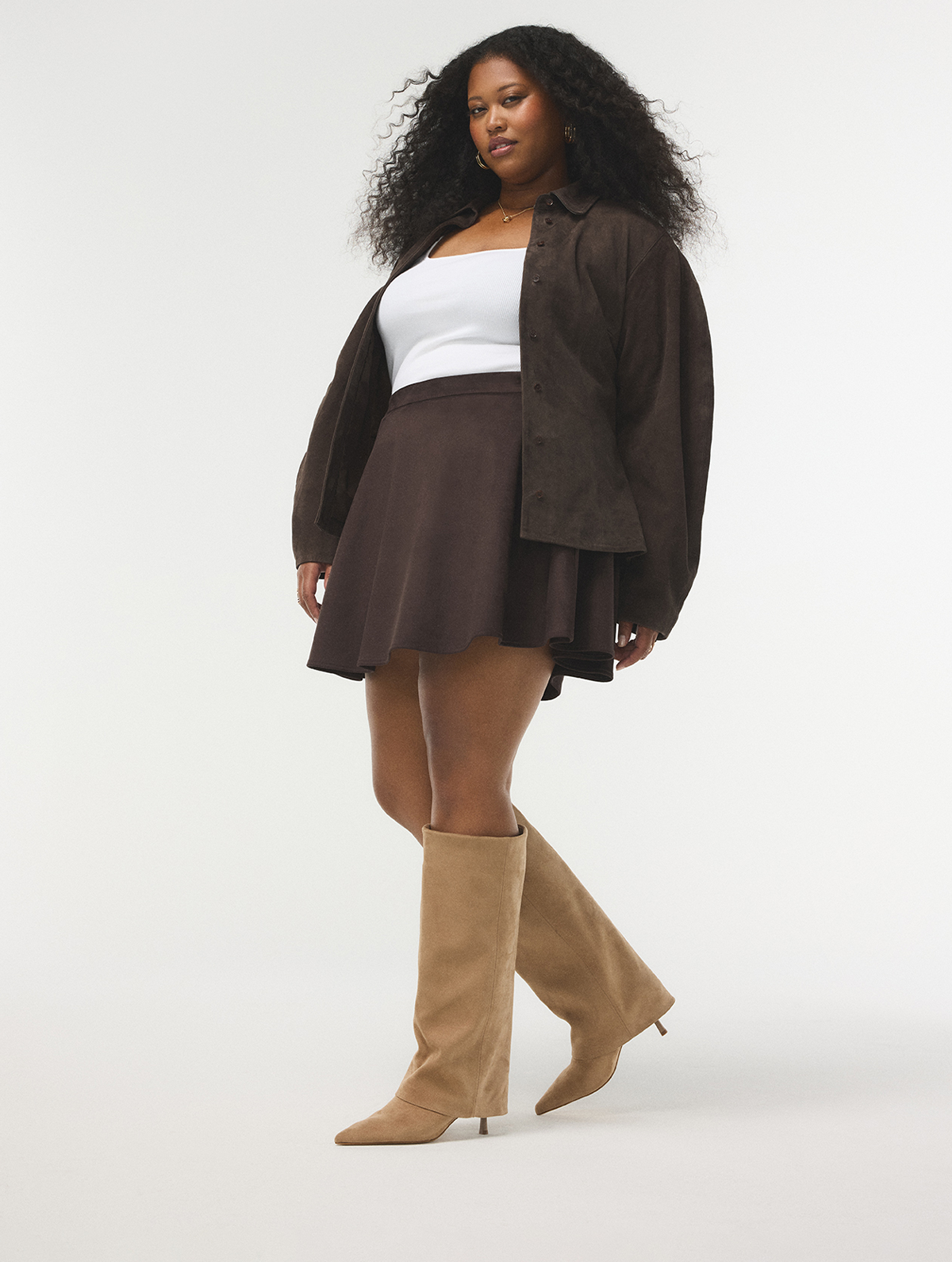 Un mannequin portant une surchemise boutonnée à effet suédé brun foncé, une minijupe plissée brun foncé, un débardeur côtelé blanc et des bottes tan à talons et à hauteur du genou, prenant la pose devant un fond de studio gris pâle sans joint visible.