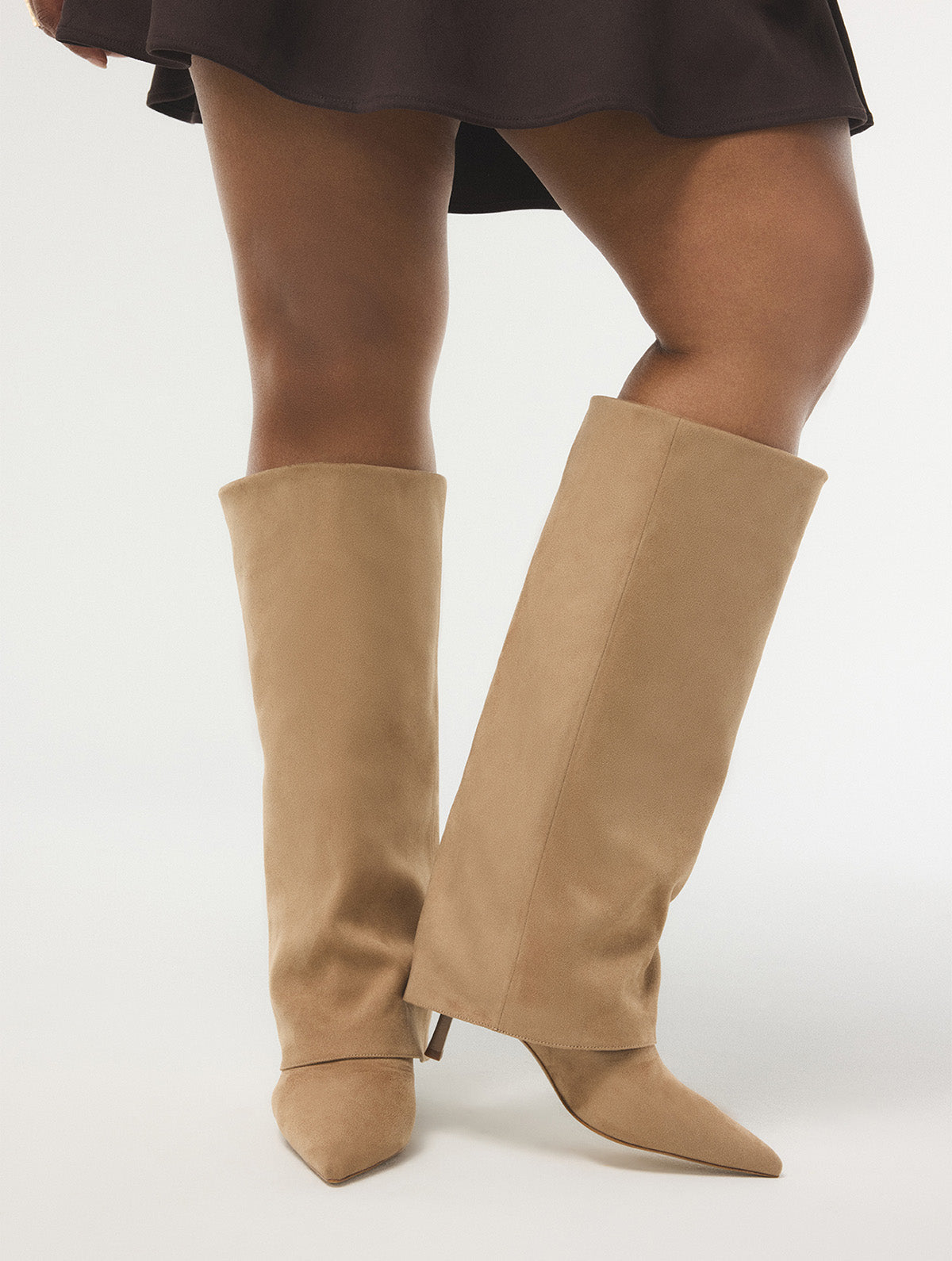 A person wearing tall knee-high pointed-toe heeled boots in a light tan suede-like finish, shown against a plain light gray studio background.