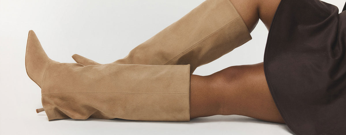 Une vue rapprochée des jambes d’une personne portant des bottes hautes en suède beige à hauteur du genou, agencées à une jupe brun foncé, sur un fond blanc épuré.